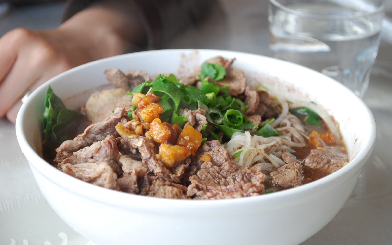 A bowl of dark noodle soup on a steel counter, adult cook's hands at the edge