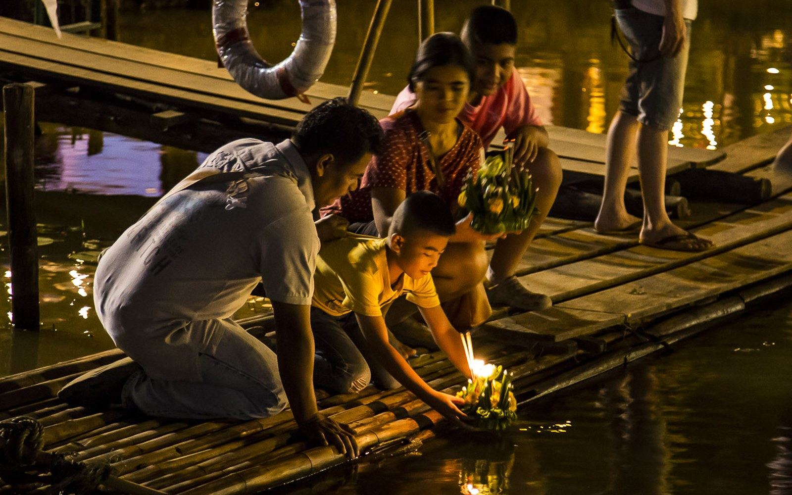 Small candle-lit floats on dark water at night, adults crouched at the bank